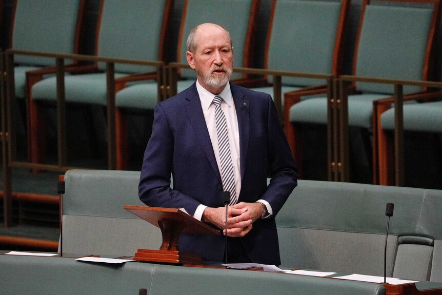 A man with a beard in a suit and tie stands with hands clasped in front of a microphone. 