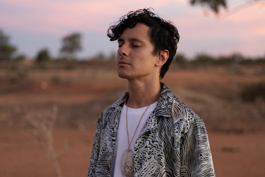 Rhyan Clapham stands with his eyes closed in front of dry scrub with red earth and a pink sunset