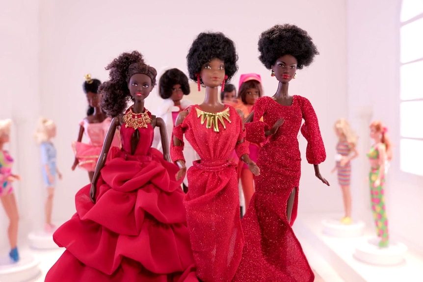 Six Black Barbies in red dresses stand in a white room with light pouring in, surrounded by white Barbies.
