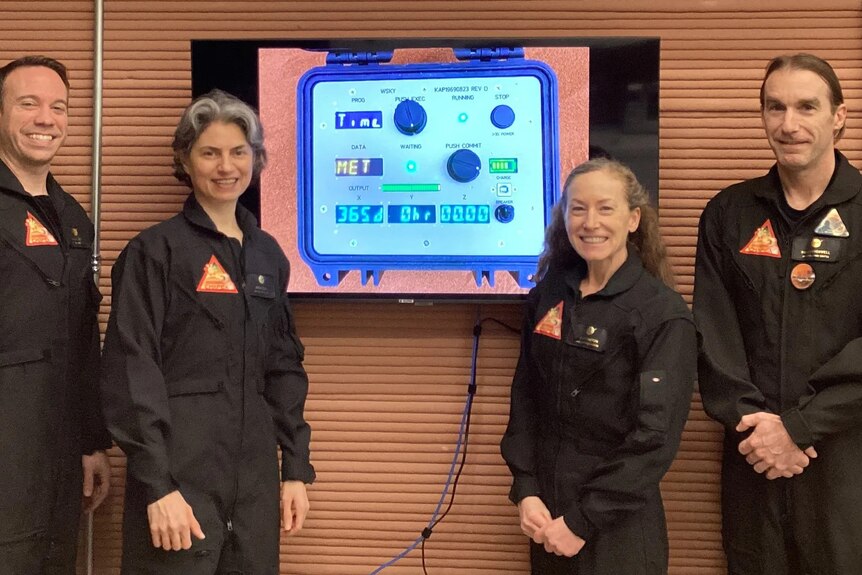 Four NASA crew members stand in front of a countdown before they enter the Mars simulation