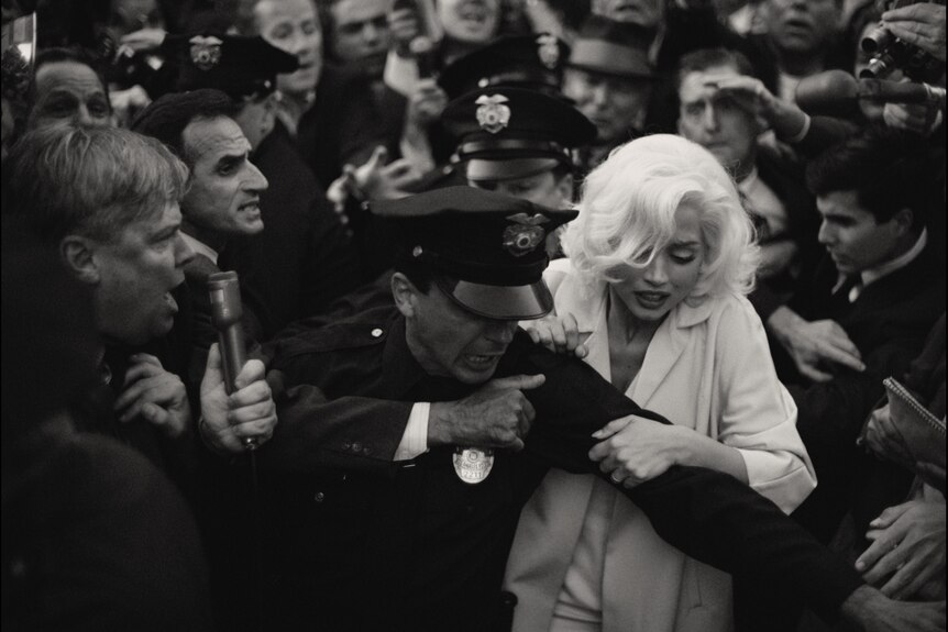 Black and white image of blonde white woman in white coat trying to squeeze through crowd of paparazzi.