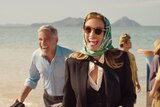 Middle-aged white man with grey hair wears blue shirt behind woman with red hair, sunglasses, head scarf and suit on a beach.