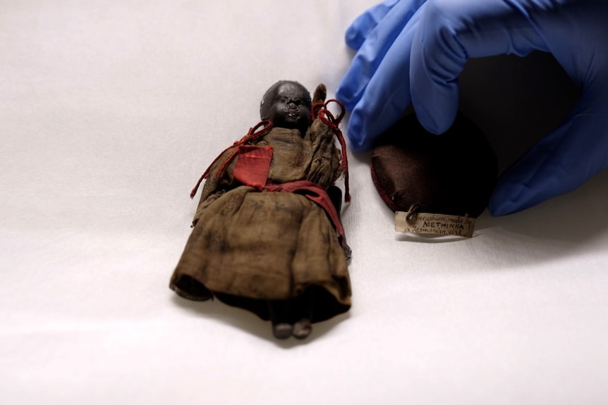 A small doll next to a pin cushion on a silk white pillow. A hand with a blue glove places the pin cushion down