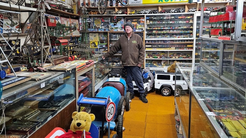 A man in a brown sweater, a black beanie and denim jeans stands in front of children's car toys. 