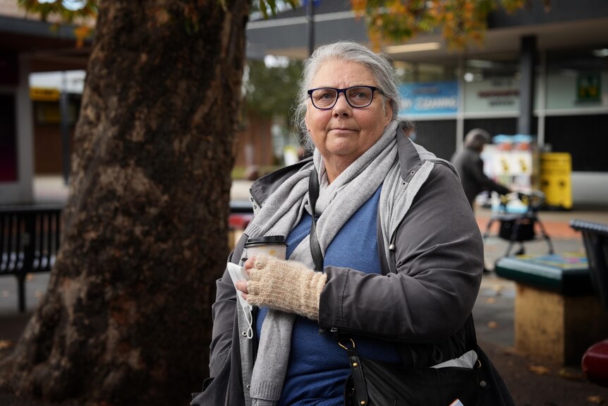 A woman holding a takeaway coffee cup and surgical mask next to a tree. 