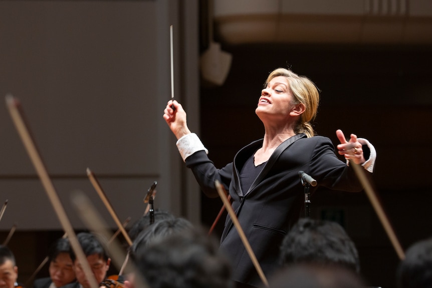 Maestro Keri-Lynn Wilson smiling widely holds her conductor's wand up high as she faces musicians before her.