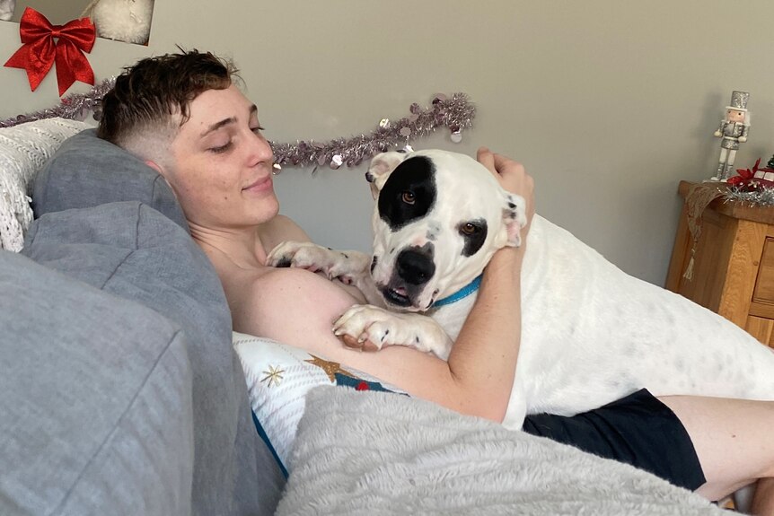 young man with short hair sits on couch whilst patting dog on his chest