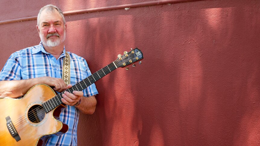 A portrait of Grant Luhrs holding his guitar leaning up against a dark red wall