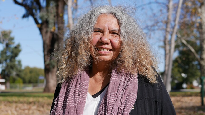 a woman standing in a park with curly hair, smiling, wearing a black jacket and purple scarf