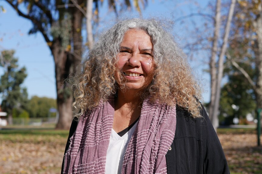 a woman standing in a park with curly hair, smiling, wearing a black jacket and purple scarf