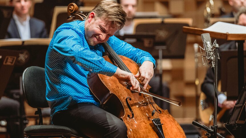 Cellist Jonathan Békés plays cello in front of a microphone. Musicians are looking on onstage in the background.