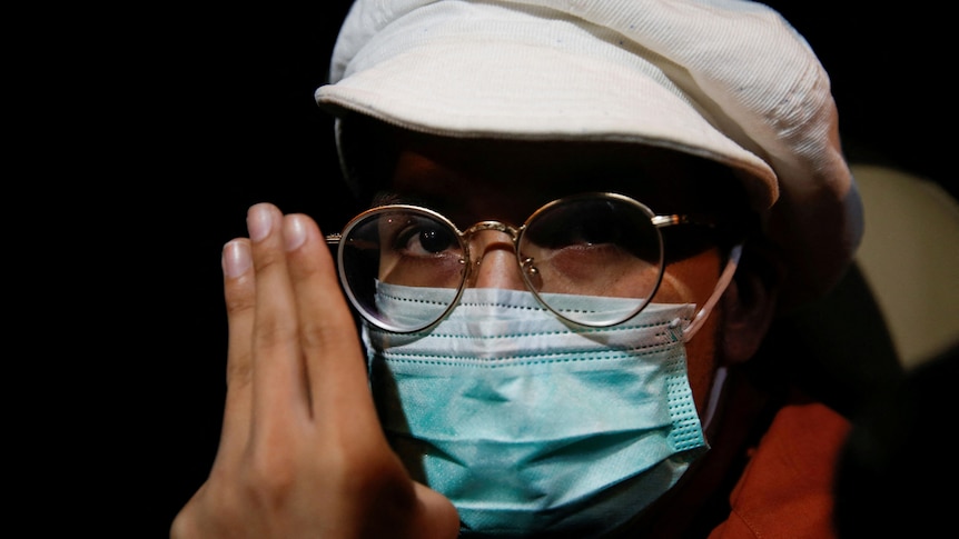 A man wears a face mask and holds his hand up to his face in a three-finger salute