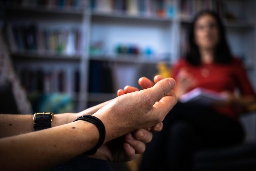 A close up of fidgeting hands. In the background, a psychologist watches on, taking notes.