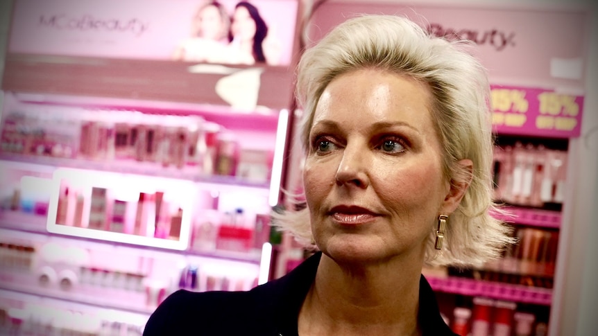 A woman with short blonde hair looks off to the side standing in front of a pink cosmetics display.