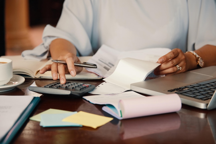 A lady sitting at a desk with lots of paperwork around her and a laptop and calculator