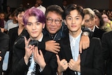 A middle-aged man in glasses smiles with his arms around two young K-pop stars, one with pink hair