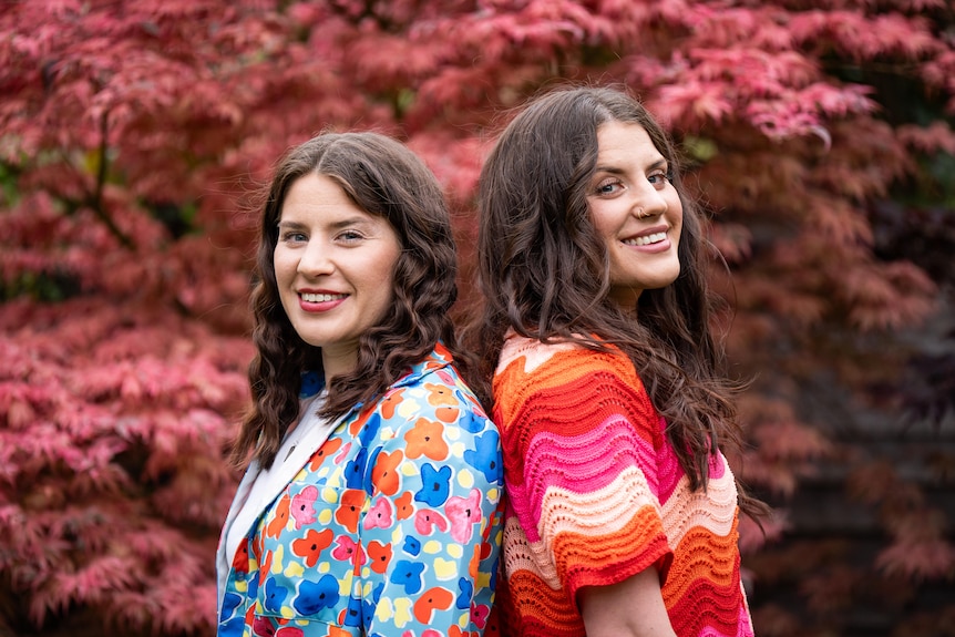 Twin sisters standing back to back smiling at the camera in very colourful tops