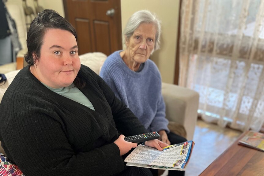 Two women, one young and the other elderly, sitting on a couch with their TV guide looking up towards the camera 