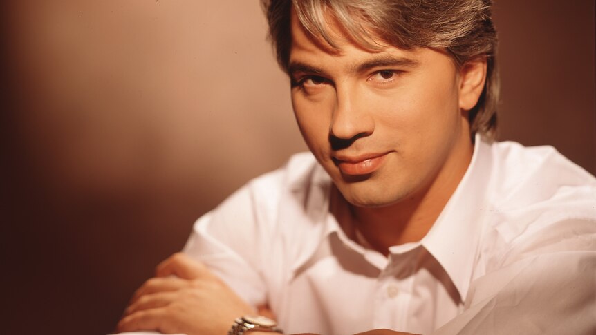 A man in a white shirt with arms crossed in front of him against a sepia background.