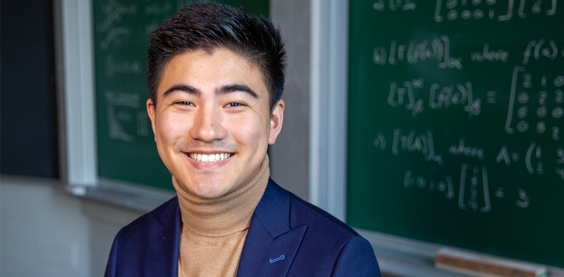 Man with short dark hair wearing a camel turtleneck under a blue blazer, sitting in front of a green chalkboard