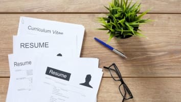 Stack of resumes laying on a table next to a pen, reading glasses, and a plant.