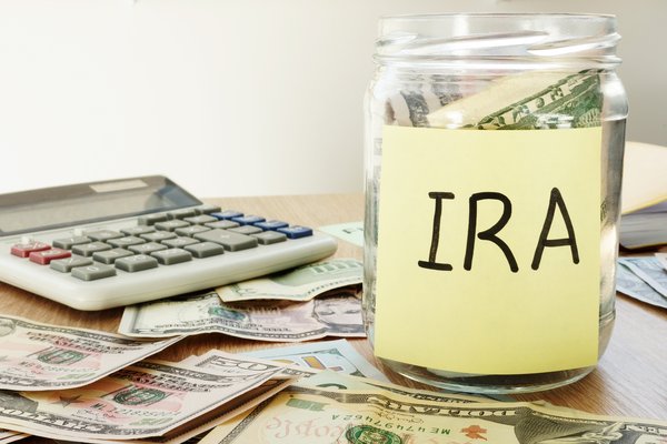 Jar of money labeled IRA sitting next to a calculator and atop various denominations of U.S. currency.