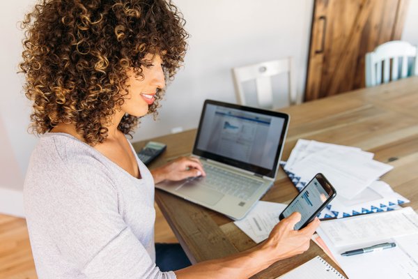 Person working on laptop and cell phone with papers at desk.