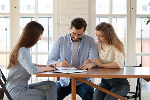 Happy young clients signing paper contract with professional broker or realtor watching.