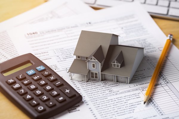 Miniature model house on top of tax forms with calculator and pencil.