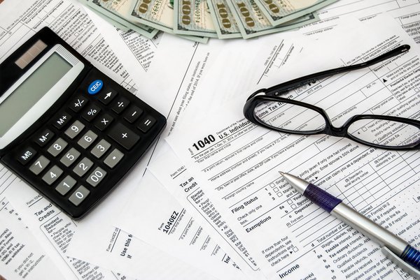 Tax forms laying scattered with a calculator, glasses, some cash, and a pen on top.