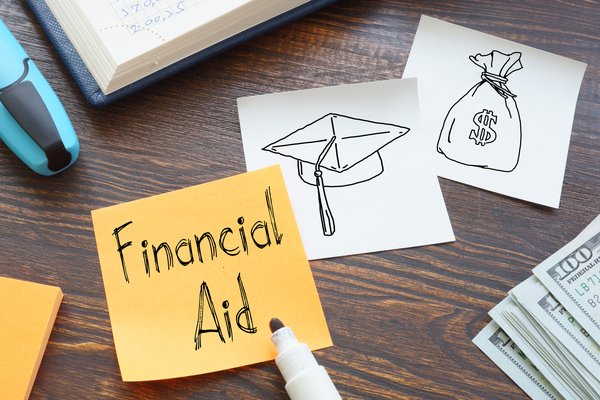 Financial aid written on a Post-It note lying on a desk next to a pile of hundred dollar bills and drawings of a cap and a bag of money.