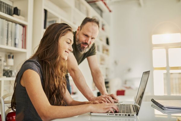 Person smiles while typing on laptop as another person stands by to help.