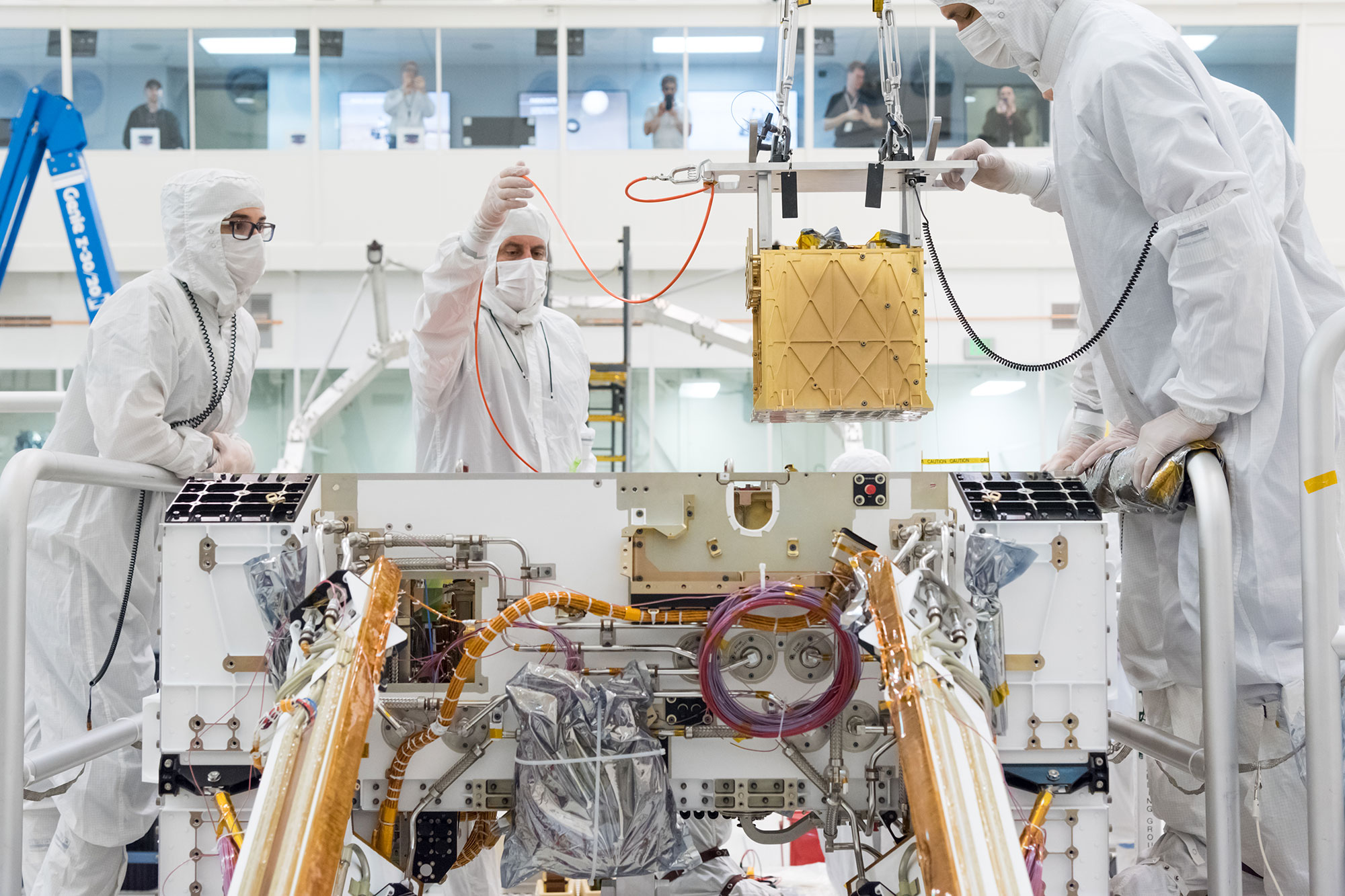 Engineers in white smocks lower a gold-colored cube into the rover