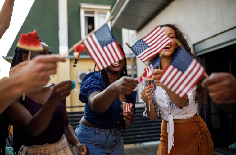 Candid shot of diverse group of joyful young people, friends, having fun, dancing and waving American flags while celebrating 4th of July on the balcony.