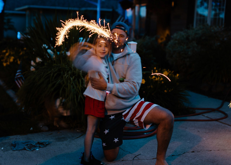 A young dad kneels down next to his toddler daughter and helps her hold a lit sparkler firework while celebrating USA Independence Day at home in the driveway on a hot summer evening.