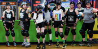 A row of roller skaters stand on the sideline of a roller skating rink