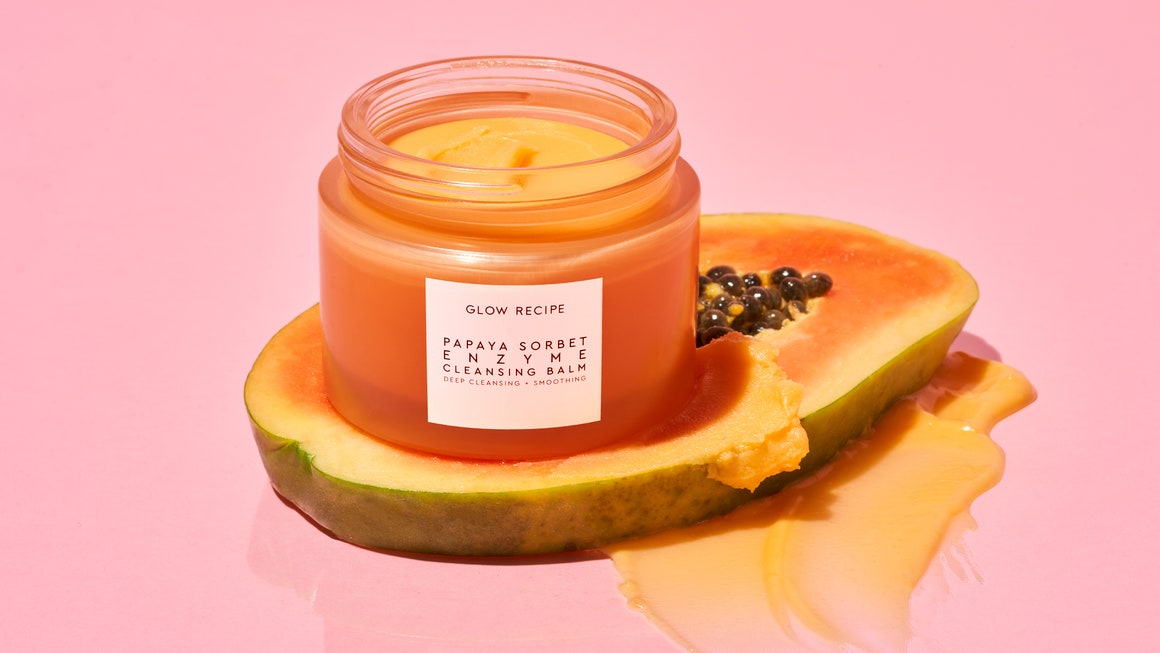 An open jar of Glow Recipe Papaya Enzyme Cleansing Balm sitting on top of a sliced piece of papaya all on a pink background