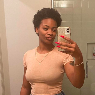 Ari Lennox in a beige fitted tshirt with short hair. She is taking a selfie in the mirror.