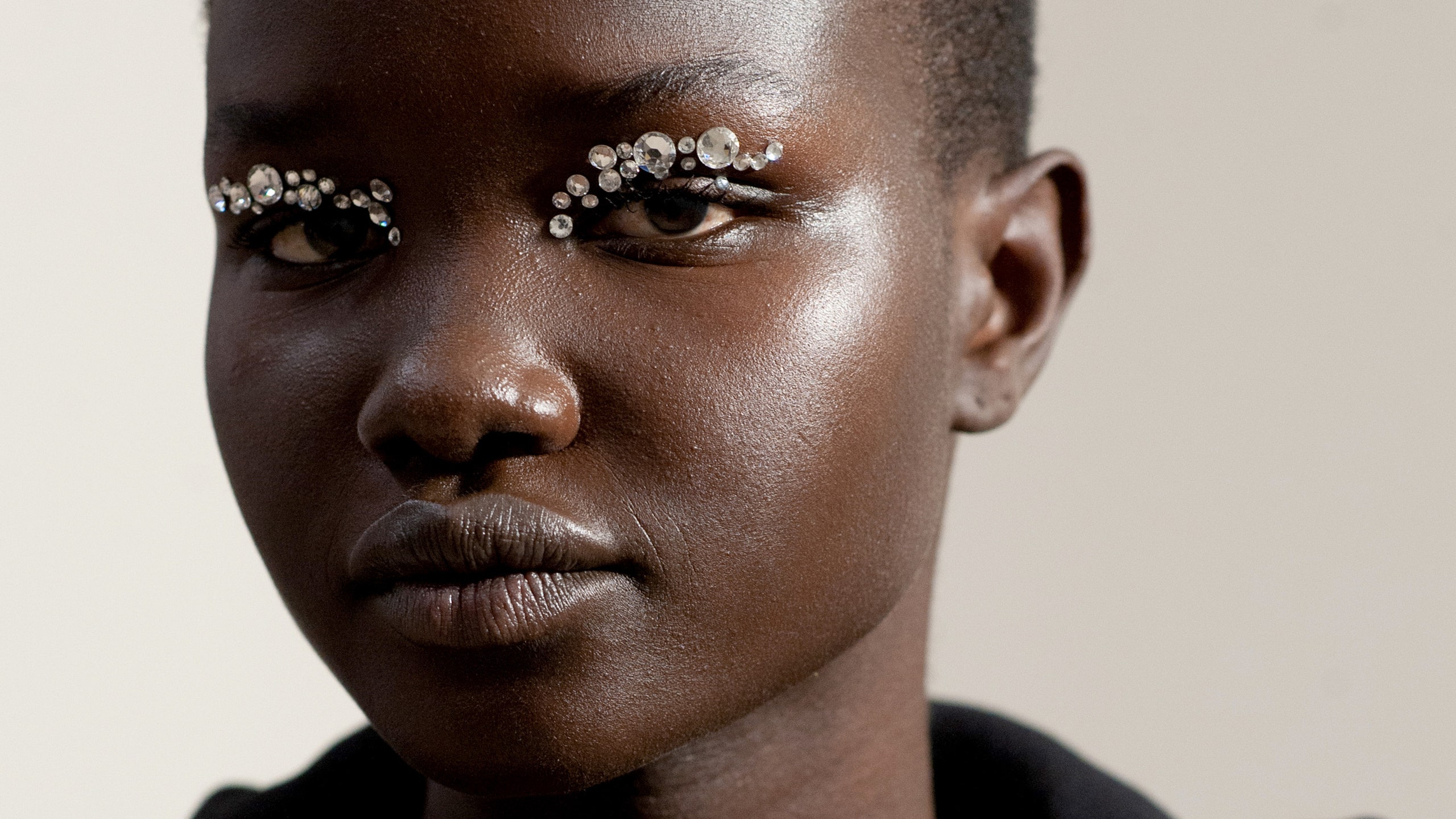 Model Akon poses backstage against a white wall with bedazzled silver eye makeup and radiant skin