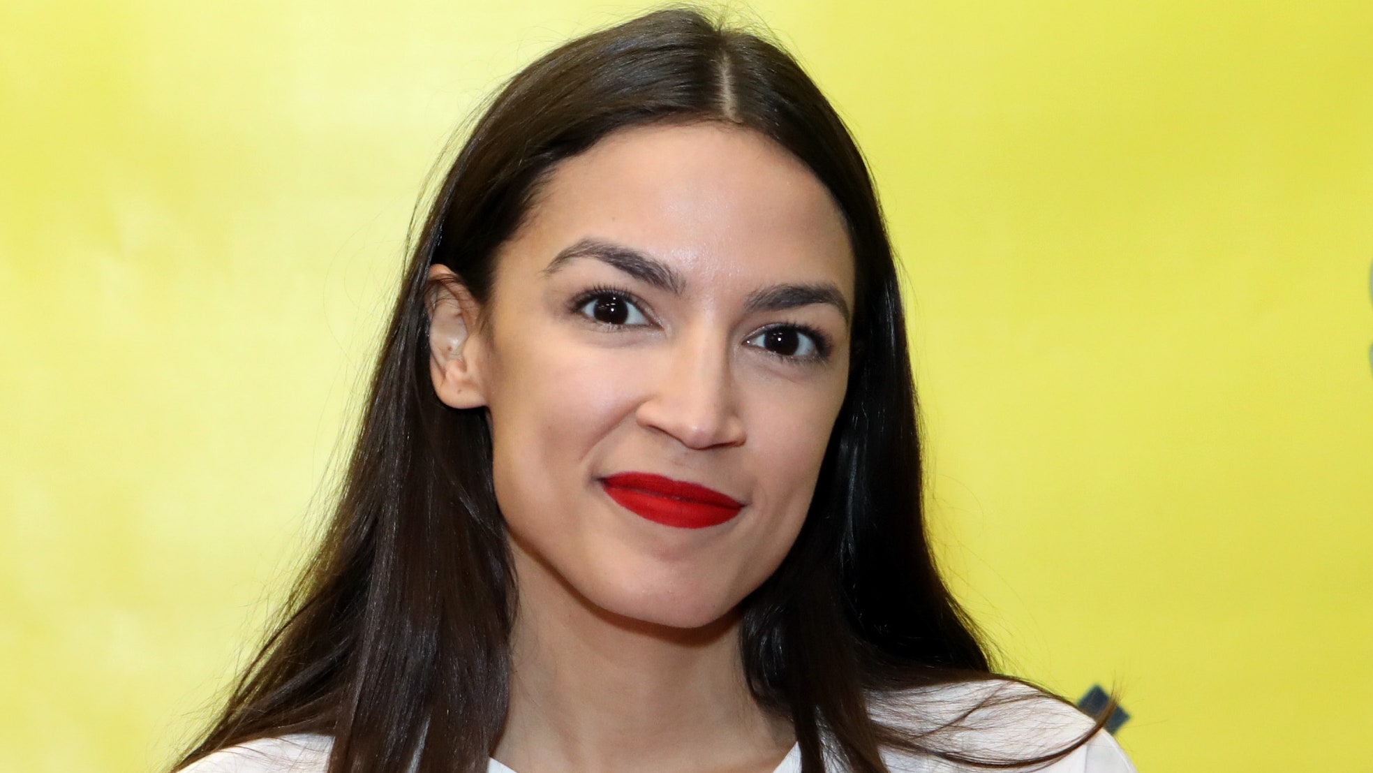 Rep. Alexandria OcasioCortez appears at an event. She wears a white shirt and red lipstick.