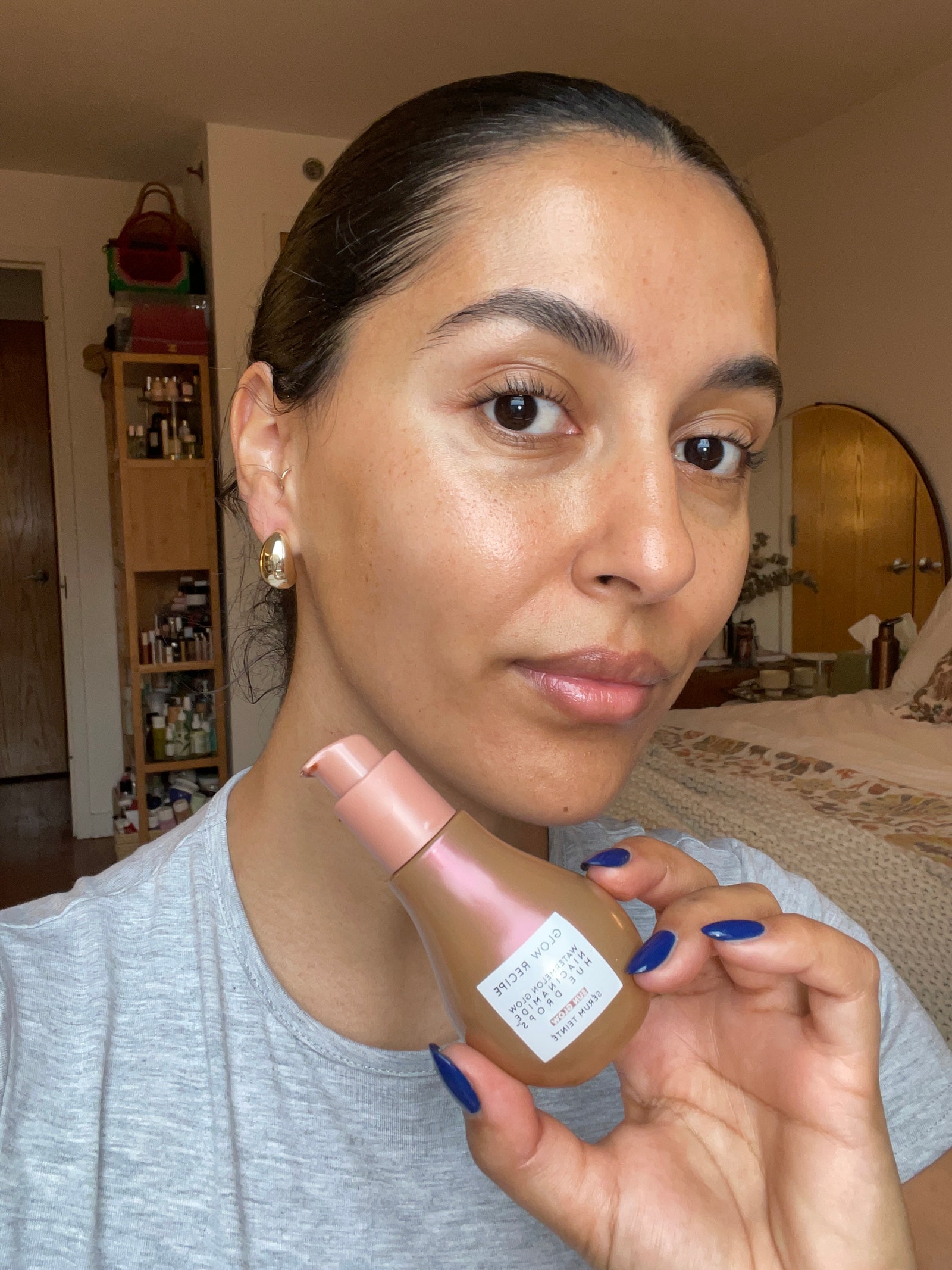 A woman with a slicked back bun smiling at camera wearing a gray tshirt holding a golden bottle of skincare serum