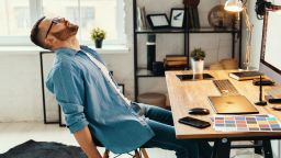 Side view of freelance male graphic designer sitting at table with large monitor and gadgets and relaxing after working hard on project