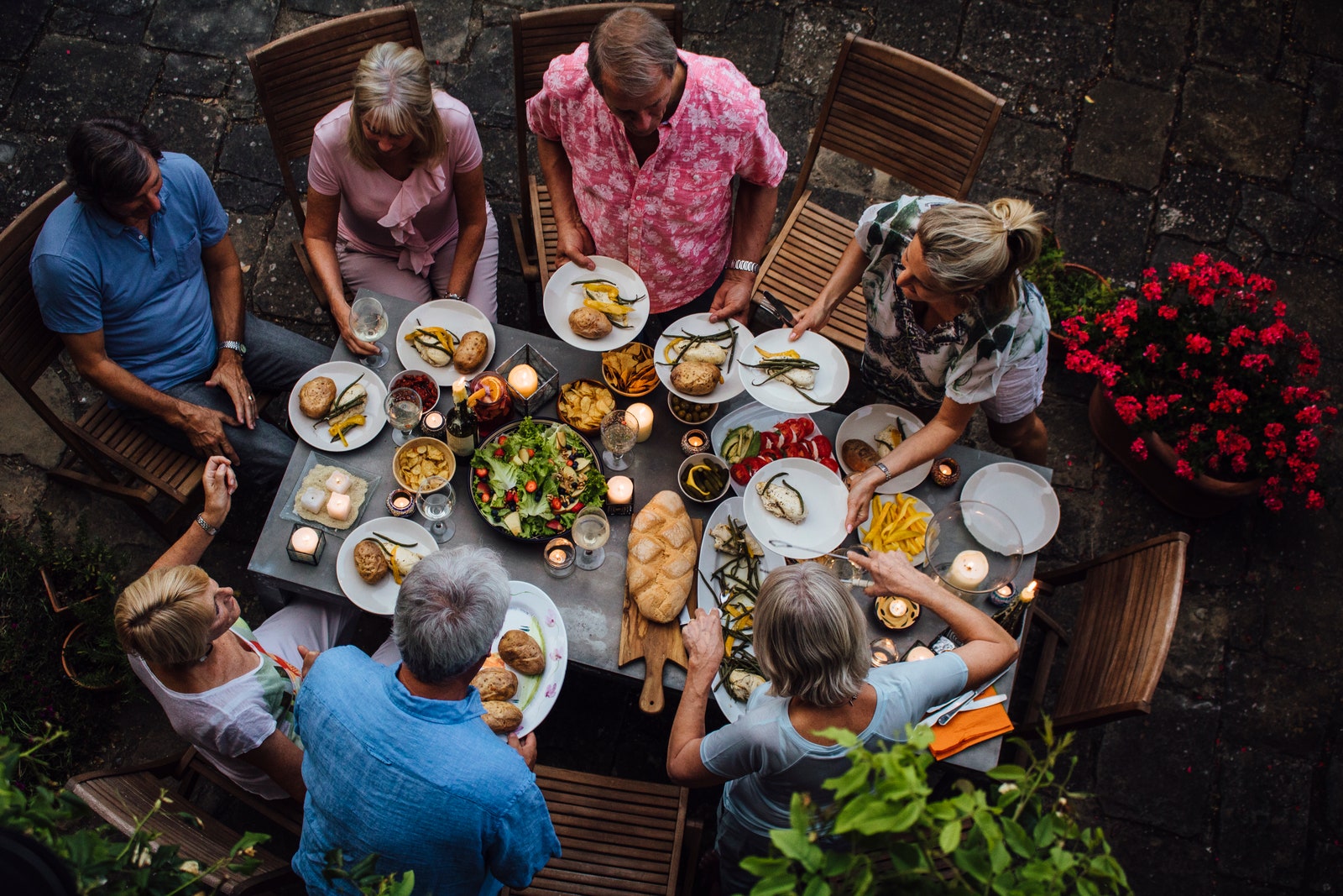 A group of mature friends are sitting around an outdoor dining table eating and drinking. They are all talking happily...