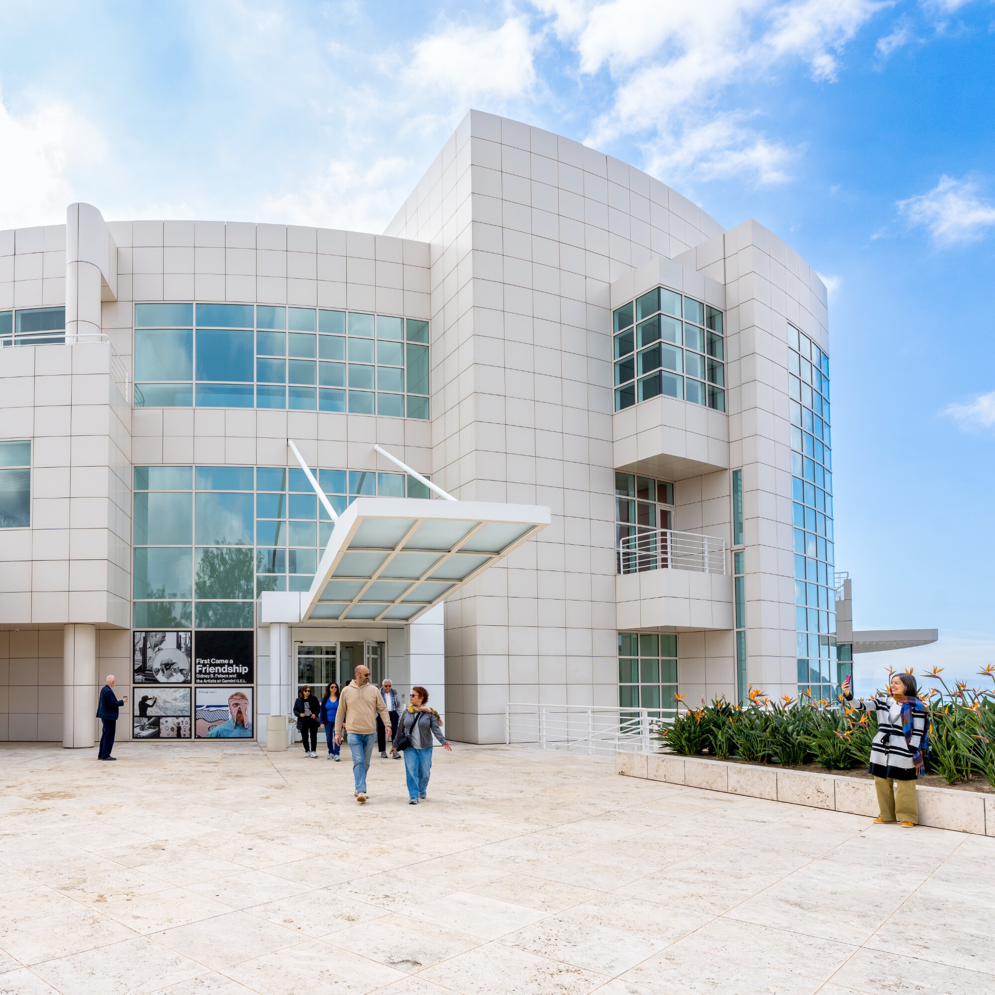 People wander in front of a circular modern building with a travertine courtyard on a sunny day