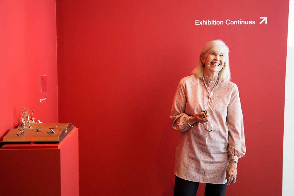 A woman in a pink shirt with a pearl necklace stands in front of a painted red wall with the words "Exhibition continues" above her head.