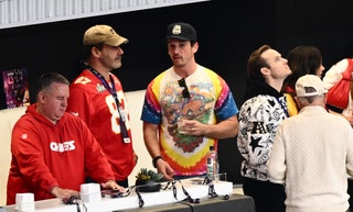 Jon Hamm wearing a Kelce Chiefs jersey and a ratty Blues cap. Miles Teller wearing a Grateful Dead tiedye tee and a logo...