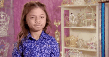 a young girl in a blue shirt is making a funny face in front of a shelf filled with crowns .