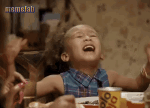 a little girl is laughing while sitting at a table with a plate of food in front of her .