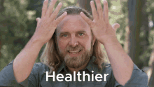 a man with long hair and a beard has his hands on his head and the word healthier is written below him