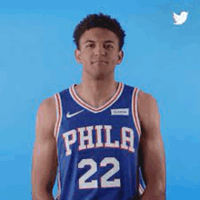 a philadelphia 76ers basketball player is standing in front of a blue background and looking at the camera .